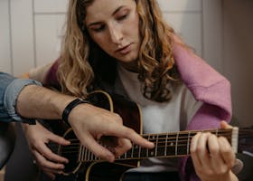 A woman is learning to play acoustic guitar with guidance indoors, focusing on finger placement.