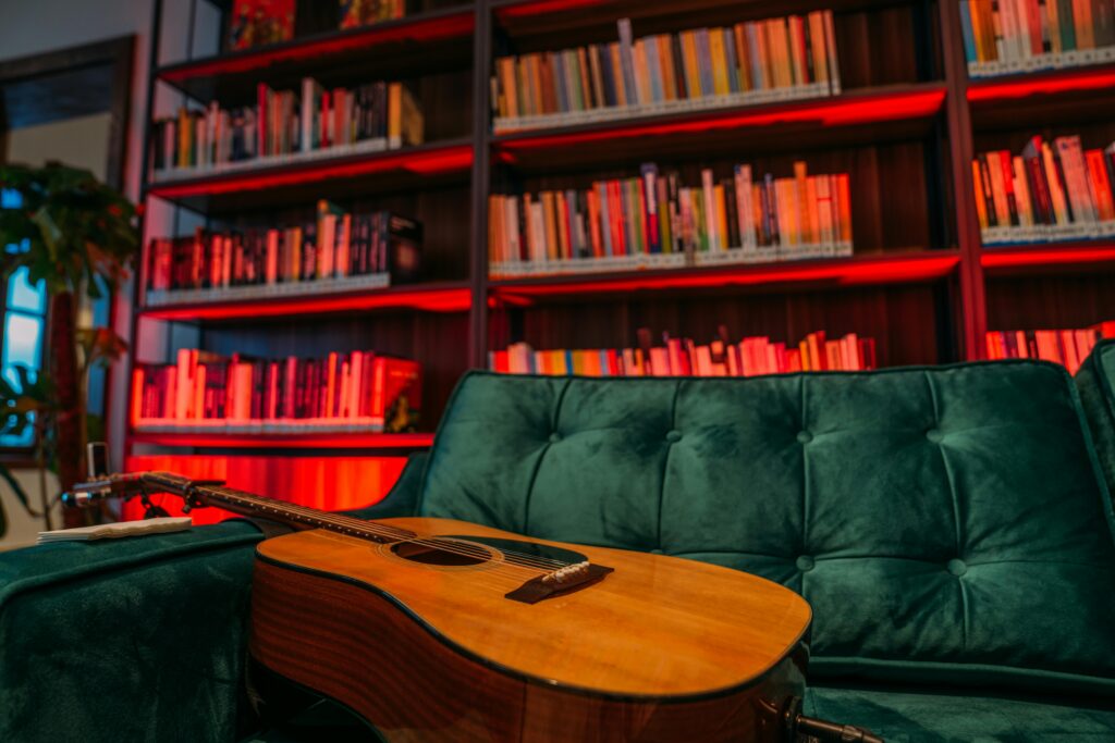 An acoustic guitar rests on a sofa in a cozy library with colorful bookshelves.